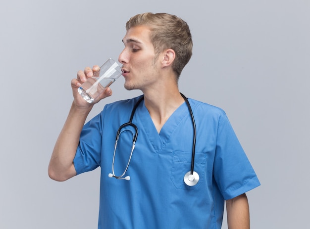 De tevreden jonge mannelijke arts die artsen eenvormig met stethoscoop draagt drinkt water dat op witte muur wordt geïsoleerd