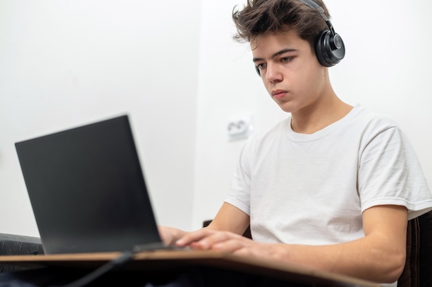 De tienerjongen gebruikt thuis laptop met hoofdtelefoons. Serieus en gefocust gezicht