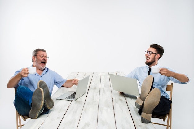 De twee glimlachende zakenlieden met benen over tafel die aan laptops op witte achtergrond werken. Zaken doen in Amerikaanse stijl