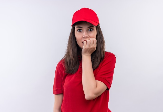 De verbaasde jonge bezorgvrouw met een rood t-shirt in een rode pet greep haar kaak op een geïsoleerde witte muur