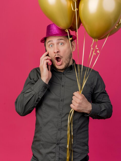 De verbaasde jonge partijkerel die roze hoed draagt die ballons houdt spreekt over telefoon die op roze wordt geïsoleerd