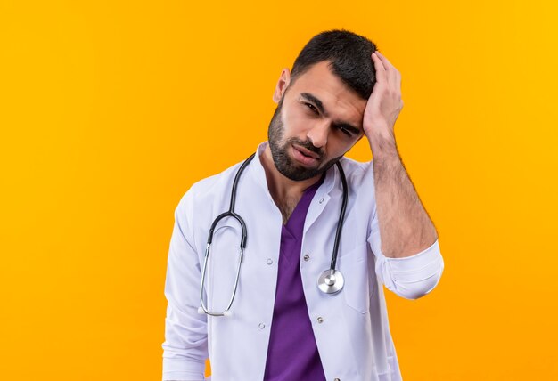 De vermoeide jonge mannelijke arts die de medische toga van de stethoscoop draagt, legde zijn hand op het hoofd op geïsoleerde gele muur