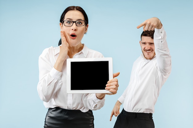 De verraste bedrijfsmens en vrouw die op een blauwe muur glimlachen en het lege scherm van laptop of tablet tonen