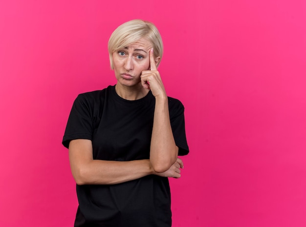 De verwarde blonde vrouw die van middelbare leeftijd naar voorzijde kijkt die denkt gebaar doet dat op roze muur wordt geïsoleerd