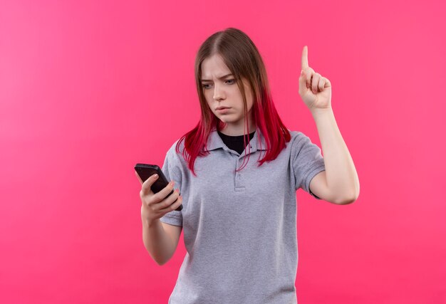De verwarde jonge mooie vrouw die grijs t-shirt draagt die telefoon in haar hand bekijkt, wijst vinger omhoog op geïsoleerde roze muur