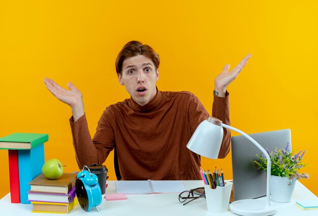 De verwarde jonge studentenjongen die aan bureau met schoolhulpmiddelen zit spreidt handen uit