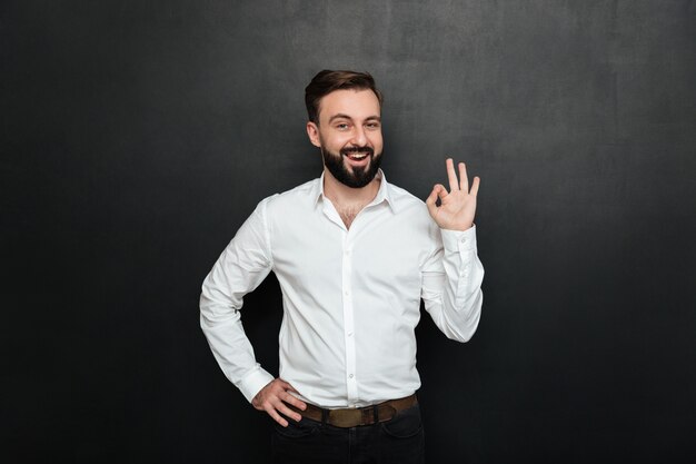 De volwassen kerel in bureau het stellen op camera, het glimlachen en het gesturing met OK teken die alles uitdrukken is in orde over donkergrijs