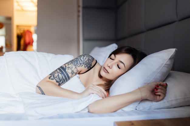 De vrij donkerbruine vrouw met tatoegering legt in het bed op het hoofdkussen in de ochtend in manier modern appartement