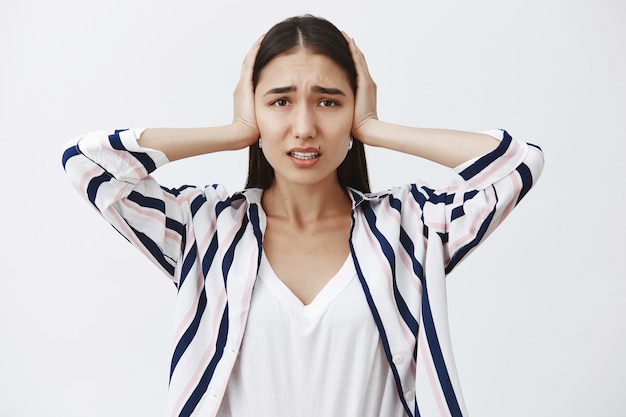 De vrouw kan de druk niet meer aan. Ontevreden sombere vrouw in gestreepte blouse, oren bedekt met handpalmen, fronsend en starend met droevige uitdrukking, isolerend van hard geluid