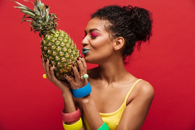 De vrouw van het mengen-ras met maniermake-up in geel overhemd die detox hebben die verse rijpe ananasholding kussen in handen geïsoleerd, over rode muur