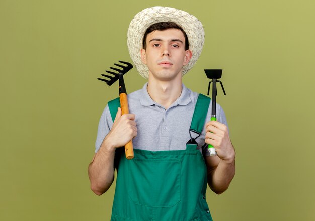 De zekere jonge mannelijke tuinman die het tuinieren hoed draagt houdt hark en schoffelhark