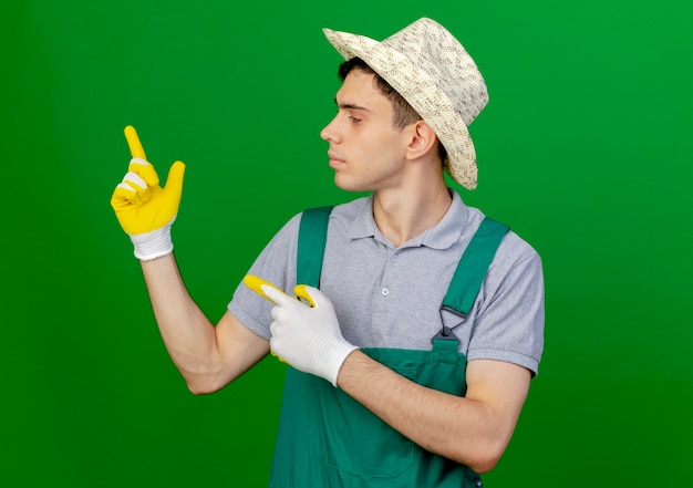 De zekere jonge mannelijke tuinman die het tuinieren hoed en handschoenen draagt kijkt en wijst naar kant