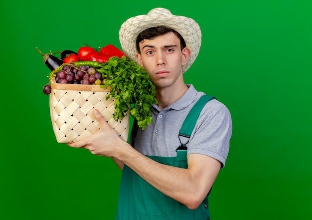 De zekere jonge mannelijke tuinman die tuinierende hoed draagt houdt plantaardige mand die op groene achtergrond met exemplaarruimte wordt geïsoleerd