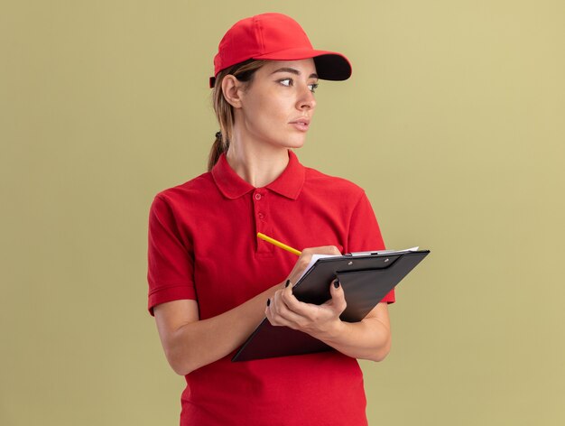 De zekere jonge mooie leveringsvrouw in uniform houdt klembord en potlood die kant bekijken die op olijfgroene muur wordt geïsoleerd