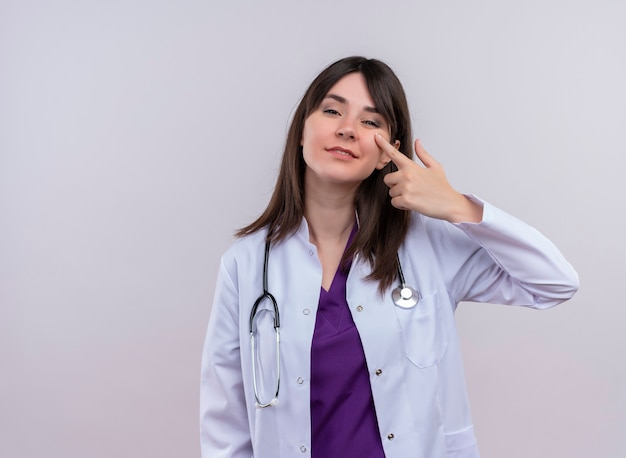 De zekere jonge vrouwelijke arts in medisch kleed met stethoscoop legt vinger op wang op geïsoleerde witte achtergrond met exemplaarruimte