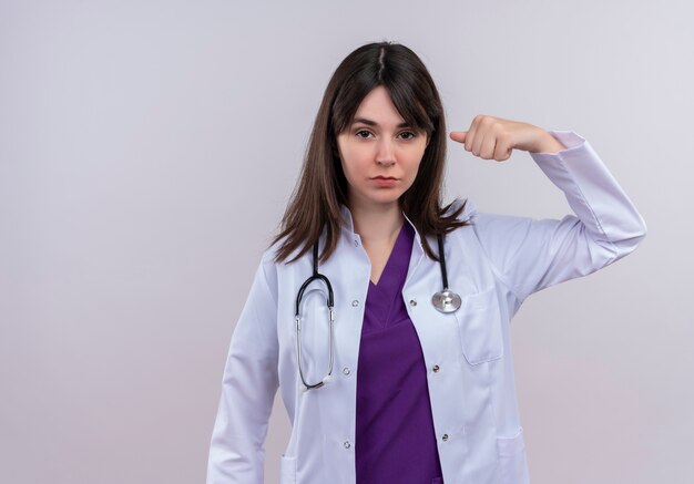 De zekere jonge vrouwelijke arts in medisch kleed met stethoscoop richt naar zichzelf op geïsoleerde witte achtergrond met exemplaarruimte