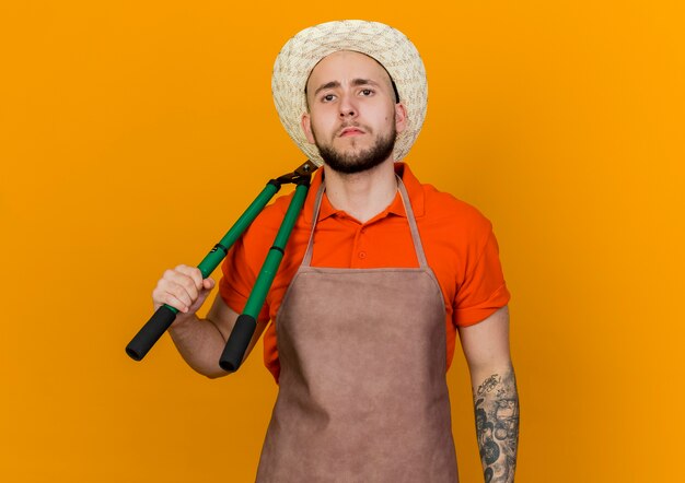 De zekere mannelijke tuinman die tuinierende hoed draagt houdt tondeuse op schouder die op oranje achtergrond met exemplaarruimte wordt geïsoleerd
