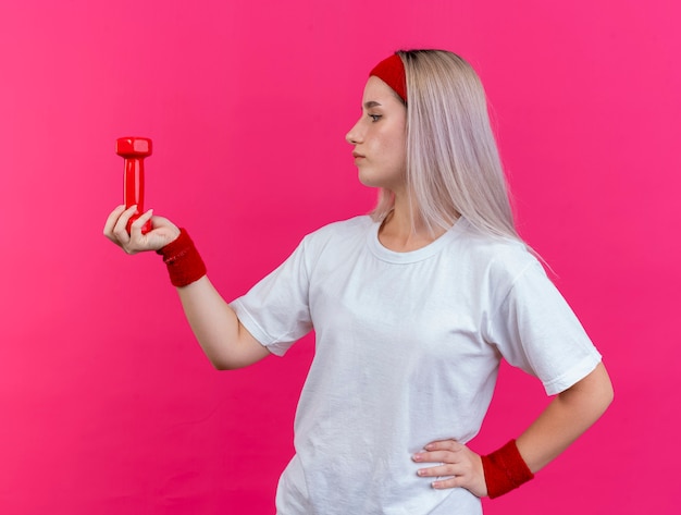 De zelfverzekerde jonge sportieve vrouw met beugels die hoofdband en polsbandjes dragen houdt en kijkt naar halter geïsoleerd op roze muur