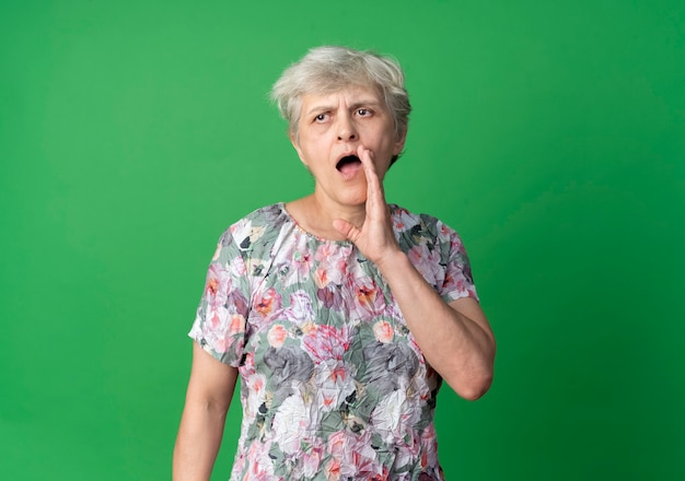 De zelfverzekerde oudere vrouw houdt de hand dicht bij de mond en doet alsof ze iemand belt die op de groene muur is geïsoleerd