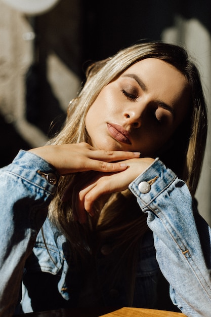 De zon schijnt over een charmante blonde in jeansjasje die bij de lijst in koffie zitten