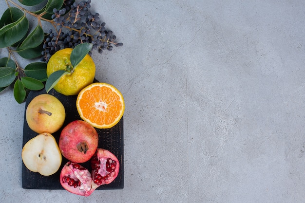 Decoratieve bladeren met gesneden en hele peren, granaatappels en mandarijnen op een zwart bord op marmeren achtergrond.