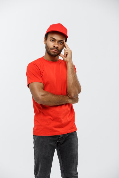 Delivery Concept - Knappe African American delivery man serieus aan iets denken. Geïsoleerd op grijze studio achtergrond. Ruimte kopiëren.