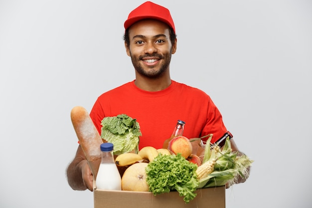 Delivery Concept - Knappe Afro-Amerikaans leveringsman met een pakket doos met kruidenierswinkel eten en drinken uit de winkel. Geïsoleerd op grijze studio achtergrond. Ruimte kopiëren.