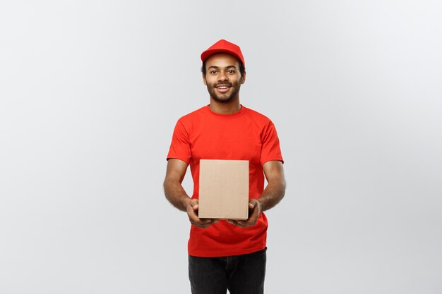 Delivery Concept - Portret van gelukkige Afro-Amerikaanse leveringsman in rode doek met een doospakket. Geïsoleerd op grijze studio achtergrond. Ruimte kopiëren.