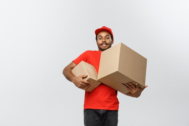 Delivery Concept - Portret van gelukkige Afro-Amerikaanse leveringsman in rode doek met een doospakket. Geïsoleerd op grijze studio achtergrond. Ruimte kopiëren.