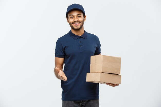 Delivery Concept - Portret van gelukkige Afro-Amerikaanse leveringsman in rode doek met een doospakket. Geïsoleerd op grijze studio achtergrond. Ruimte kopiëren.