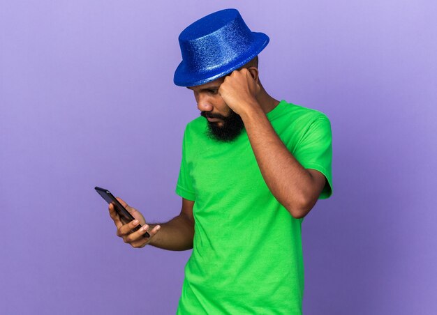 Denken aan een jonge Afro-Amerikaanse man met een feesthoed die vasthoudt en naar een telefoon kijkt die op een blauwe muur is geïsoleerd