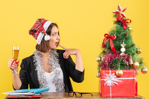 Denken charmante dame in pak met kerstman hoed en nieuwe jaar decoraties met wijn in het kantoor op geel geïsoleerd