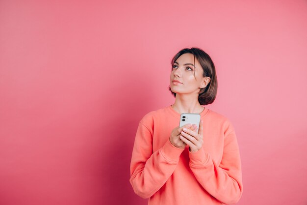 Denken dromen jonge mooie vrouw poseren geïsoleerd over roze muur achtergrond met behulp van mobiele telefoon