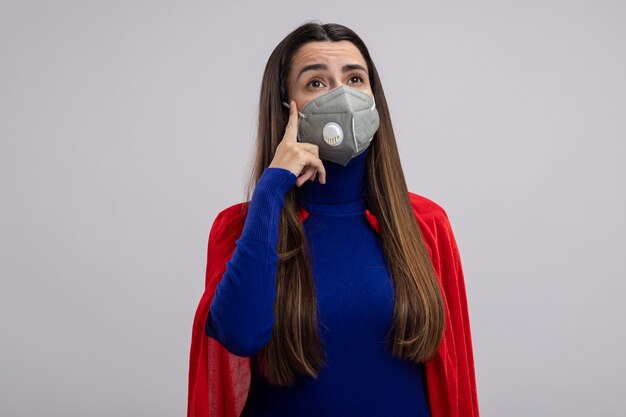 Denken jong superheld meisje kijken kant dragen medische masker vinger zetten tempel geïsoleerd op een witte achtergrond