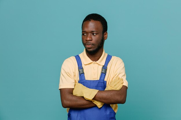 Denken kruisende handen jonge Afro-Amerikaanse schonere man in uniform met handschoenen geïsoleerd op blauwe achtergrond