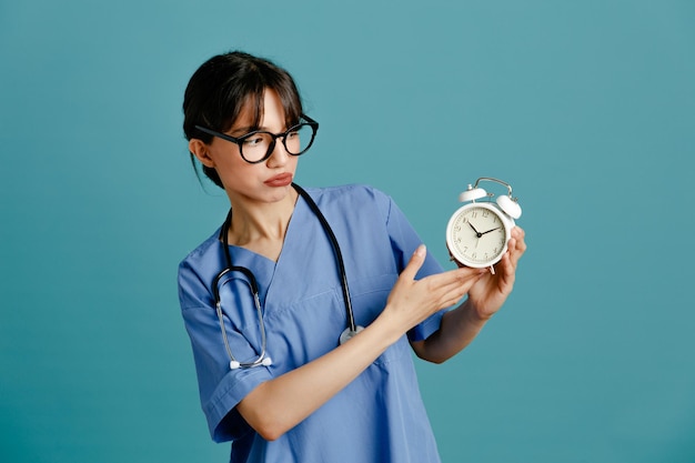 Denken met wekker jonge vrouwelijke arts dragen uniform fith stethoscoop geïsoleerd op blauwe achtergrond