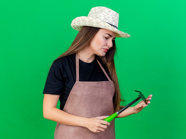 Denken mooi tuinman meisje in uniform dragen tuinieren hoed bedrijf en kijken naar schoffel hark geïsoleerd op groene achtergrond