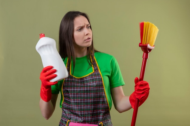 Denken schoonmakend jong meisje die uniform in rode handschoenen dragen die reinigingsmiddel houden die dweil op haar hand op geïsoleerde groene muur bekijken