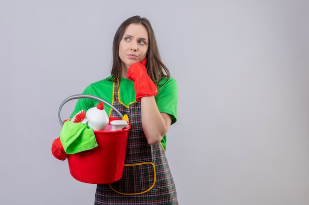 Denken schoonmakend jong meisje die uniform in rode handschoenen dragen die schoonmakende hulpmiddelen houden legde haar hand op wang op geïsoleerde witte muur