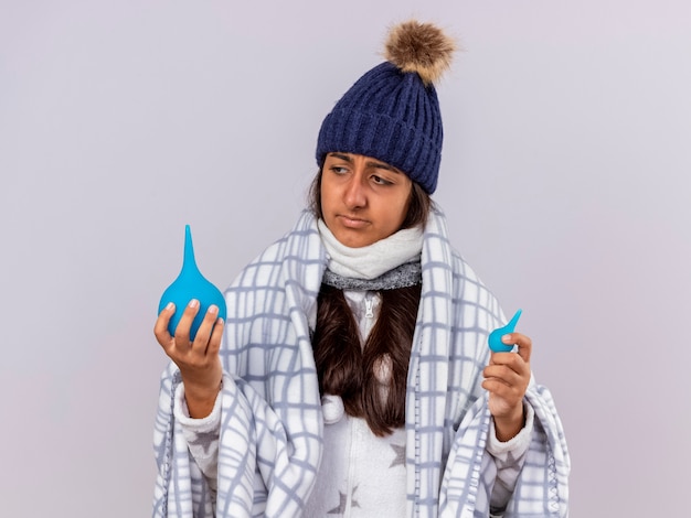 Denken ziek meisje dragen winter hoed met sjaal verpakt in geruite bedrijf en klysma te kijken