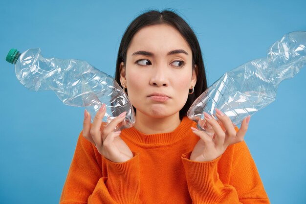 Denkend boos meisje toont twee flessen die plastic recyclen, gaat groen herbruikbaar afval blauwe achtergrond