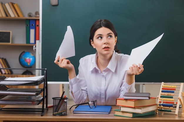 denkend kijkend naar de zijkant jonge vrouwelijke scheurpapierleraar zit aan bureau met schoolhulpmiddelen in de klas