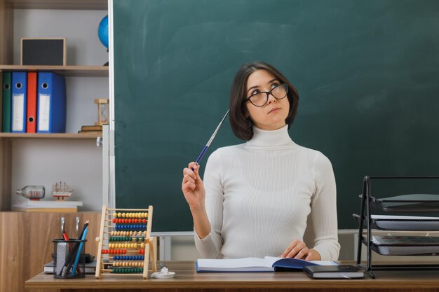 denkend kijkend naar jonge vrouwelijke leraar met een bril die de aanwijzer op het hoofd zet, zittend aan een bureau met schoolhulpmiddelen in de klas