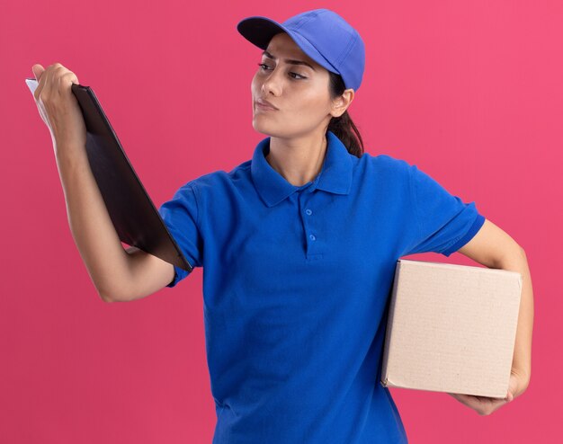 Denkende jong leveringsmeisje die uniform met de doos van de glbholding dragen en klembord in haar hand bekijken dat op roze muur wordt geïsoleerd