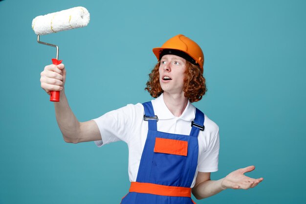 Denkende jonge bouwer man in uniform houden en kijken naar rolborstel geïsoleerd op blauwe achtergrond