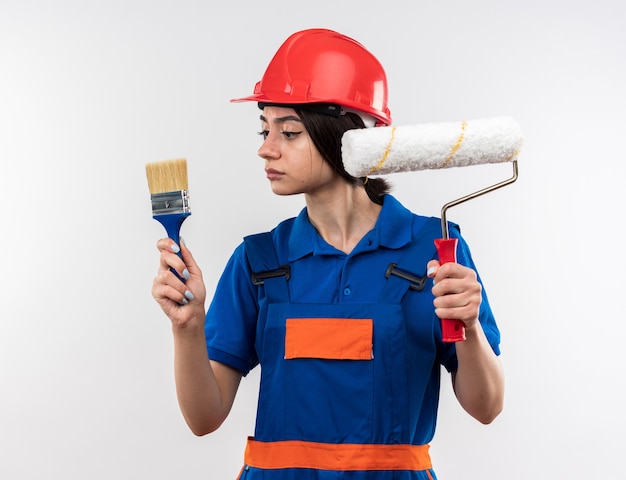 Denkende jonge bouwer vrouw in uniform houden en kijken naar rolborstel met kwast geïsoleerd op een witte muur