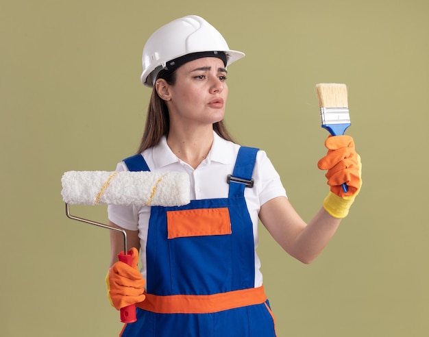 Denkende jonge bouwvrouw in uniform en handschoenen met rolborstel en kijken naar verfborstel in haar hand geïsoleerd op olijfgroene muur