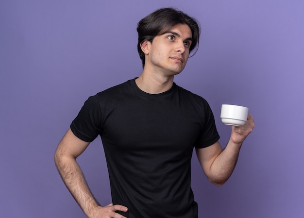 Denkende jonge knappe kerel die zwarte t-shirt draagt die kop van koffie houdt die hand op heup zet die op purpere muur wordt geïsoleerd