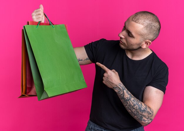 Denkende jonge knappe man met een zwart t-shirt in zijn hand en wijst naar papieren zakken geïsoleerd op roze achtergrond