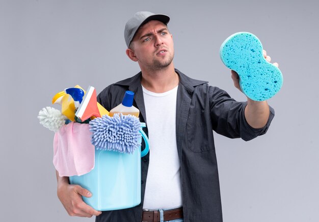 Denkende jonge knappe schoonmaakster die t-shirt en pet draagt die emmer met schoonmaakgereedschap houdt en spons in zijn hand bekijkt die op witte muur wordt geïsoleerd